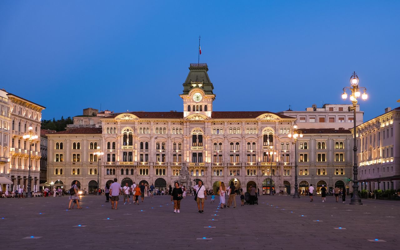 Evening view of Unity of Italy Square in Trieste, with people enjoying the illuminated architecture.