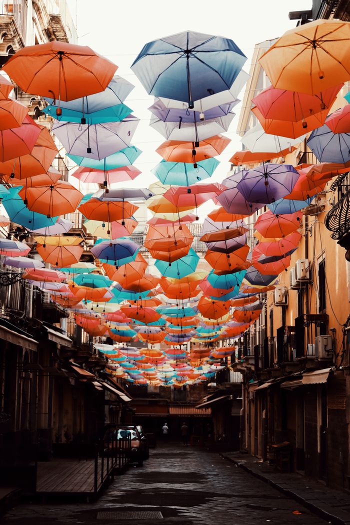 Vibrant umbrellas hang over a charming alley in Catania, Sicily, Italy, creating a lively urban scene.