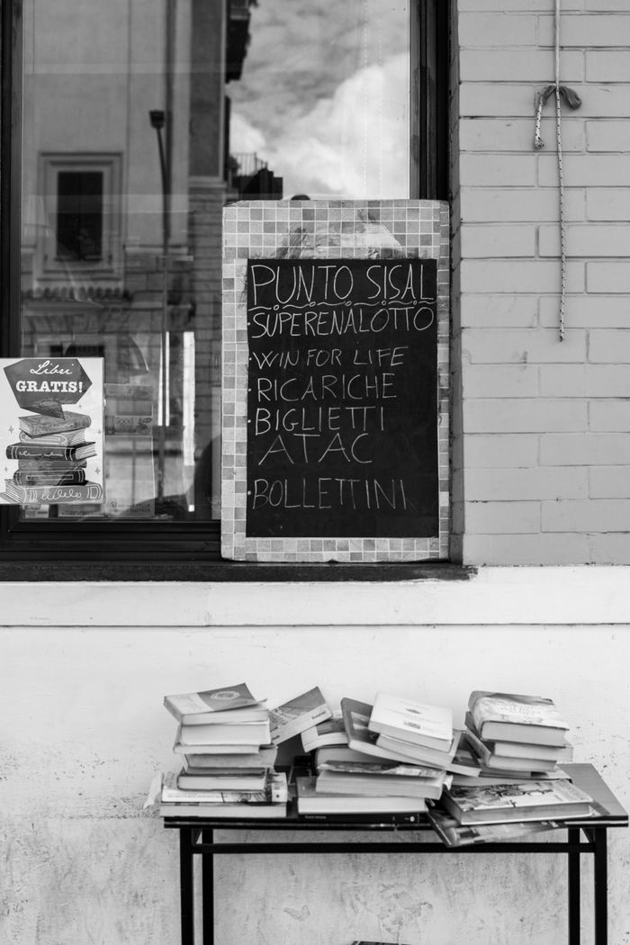 Monochrome image of a blackboard on a wall beside a window and stacked books on a table.