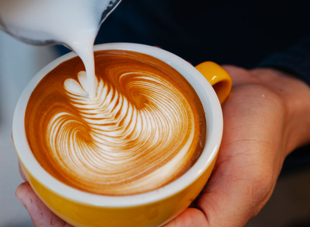 Unrecognizable professional barista pouring milk from pitcher into yellow cup with latte with creative pattern while working in coffee house